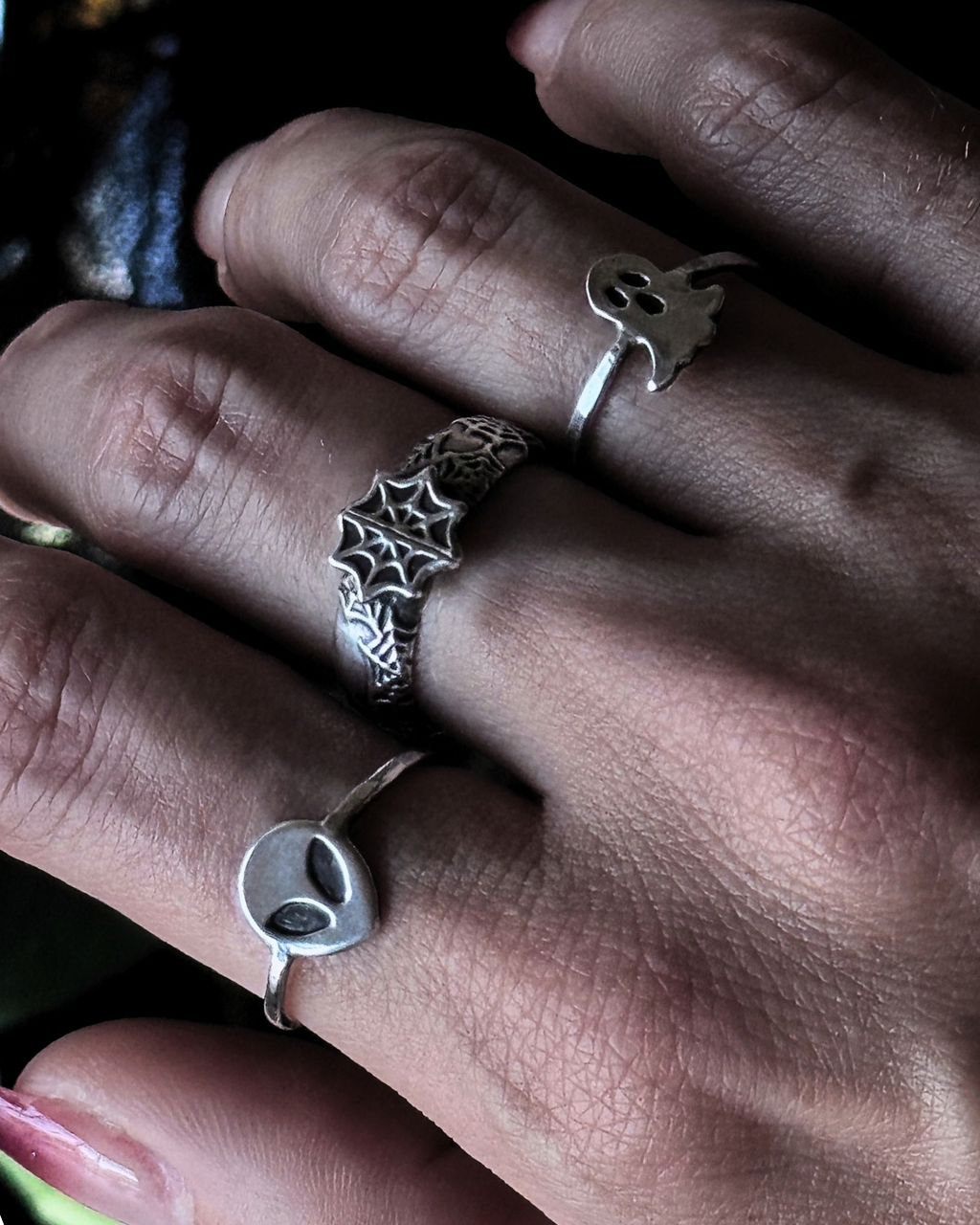 A close-up of a hand wearing three silver rings: a spiderweb ring on the middle finger, a small ghost ring on the ring finger, and an alien head ring on the pinky finger.