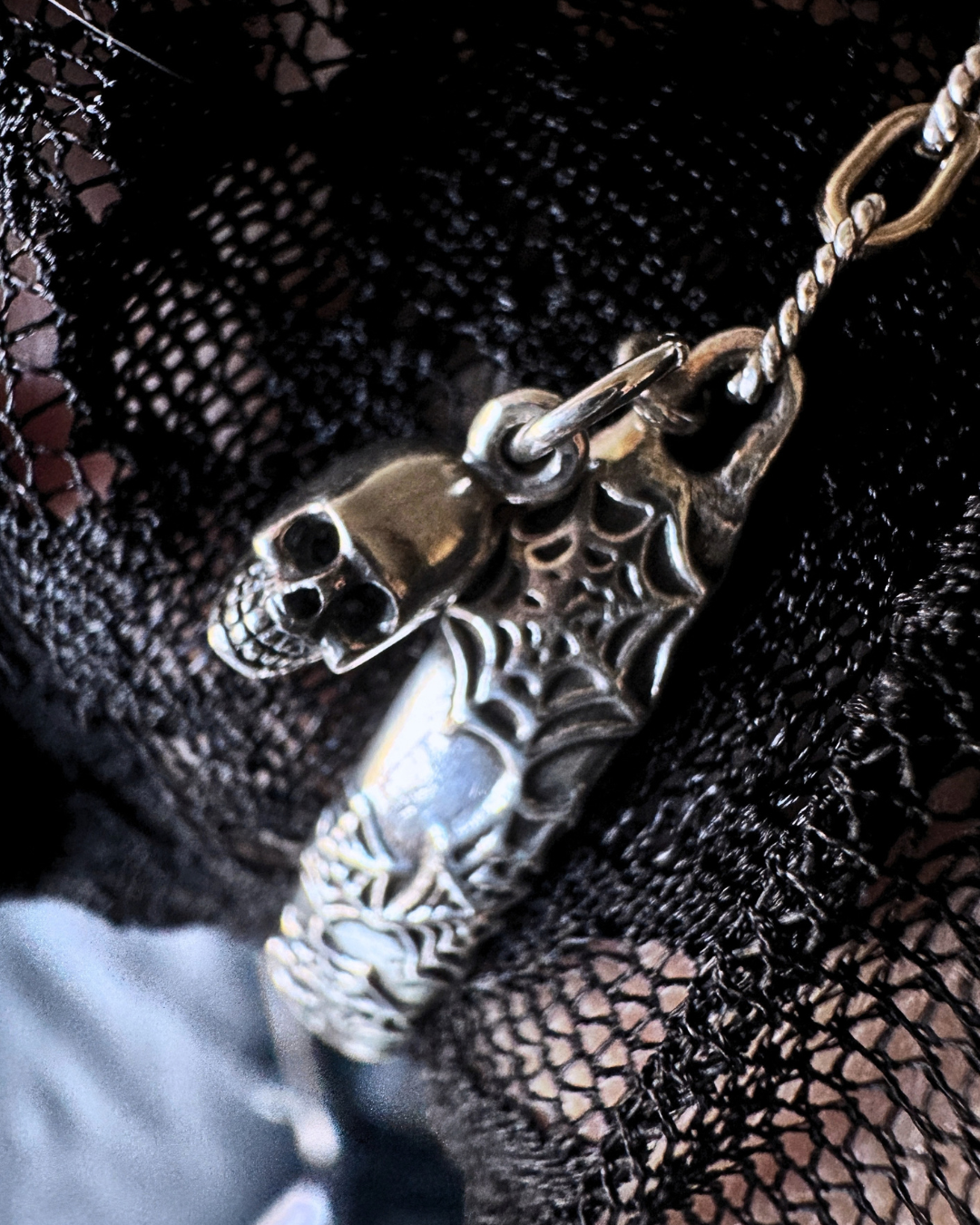 A close-up of a silver-toned metal pendant featuring a skull and a spiderweb design, resting on black lace fabric.