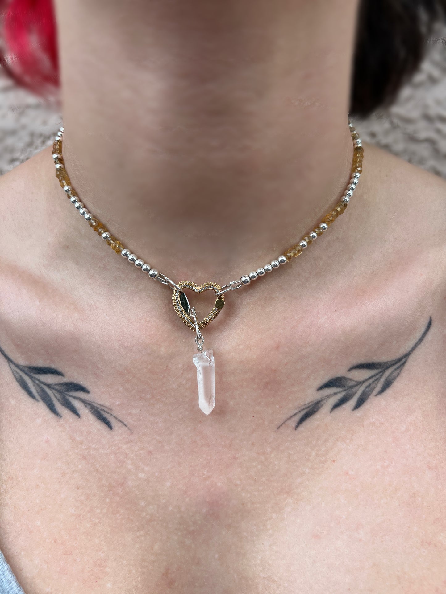 A close-up image of a sterling silver necklace with a heart-shaped clear quartz crystal and hematite accents, displayed on a person.