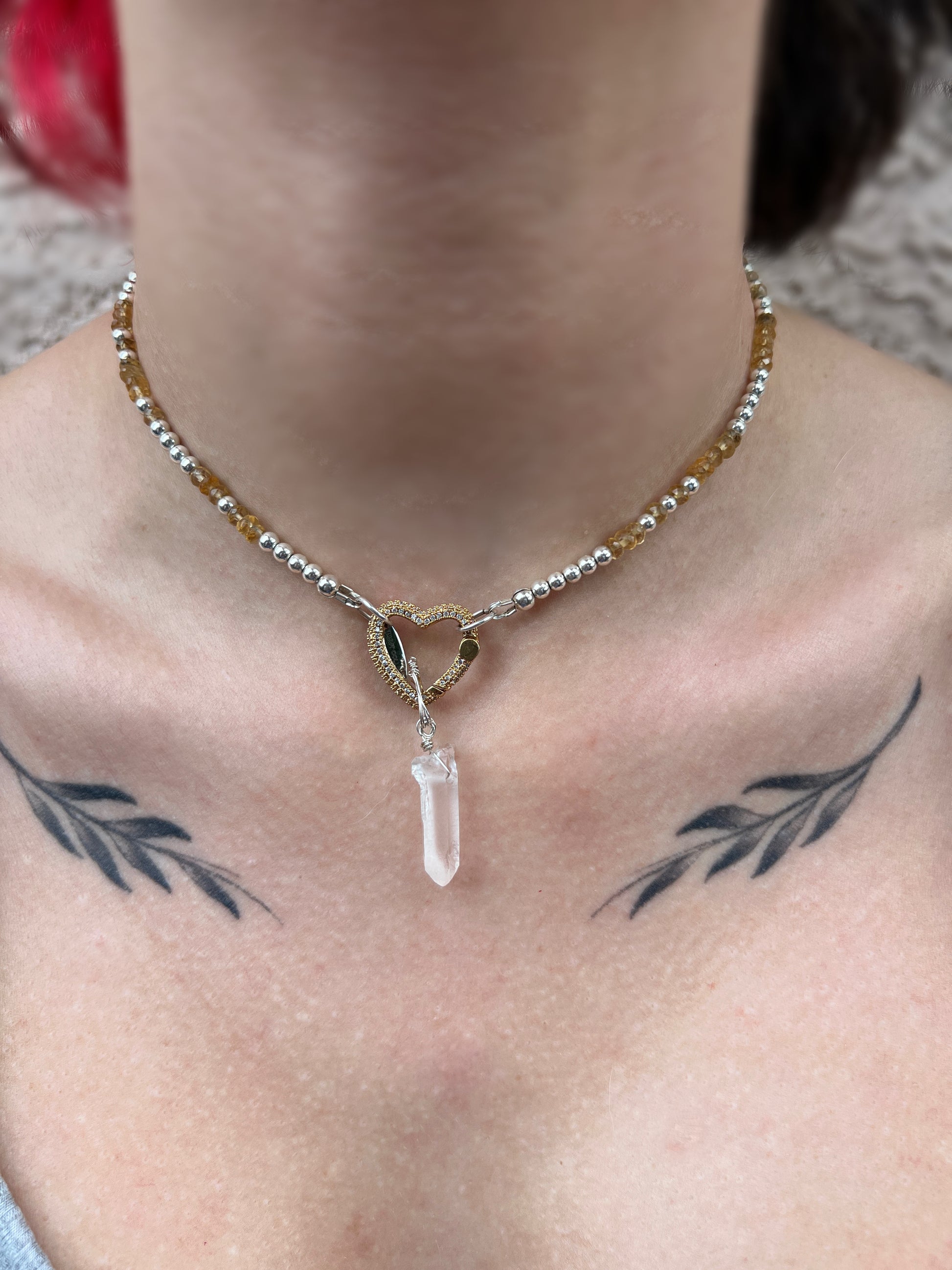 A close-up image of a sterling silver necklace with a heart-shaped clear quartz crystal and hematite accents, displayed on a person.