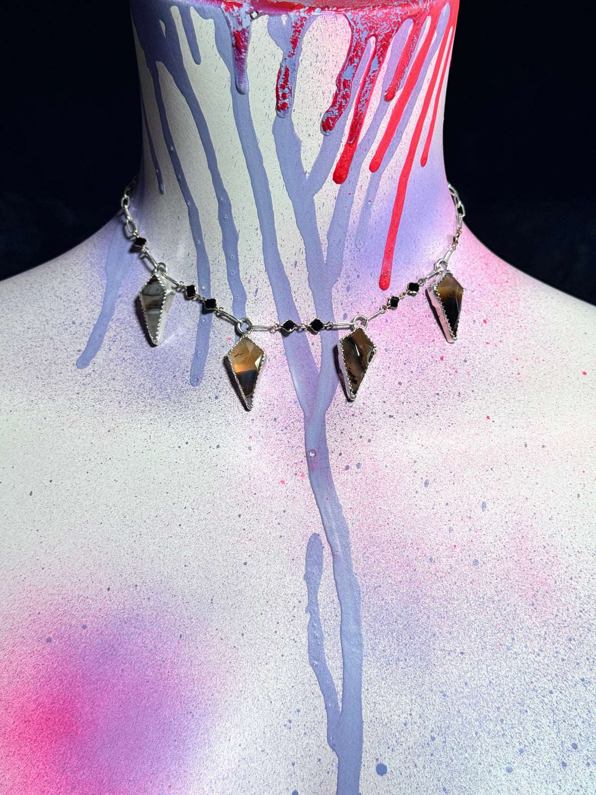 A silver-toned choker necklace with five triangular gold and brown gemstone pendants and small black beads, displayed on a painted white mannequin neck with red and blue paint drips against a black background.