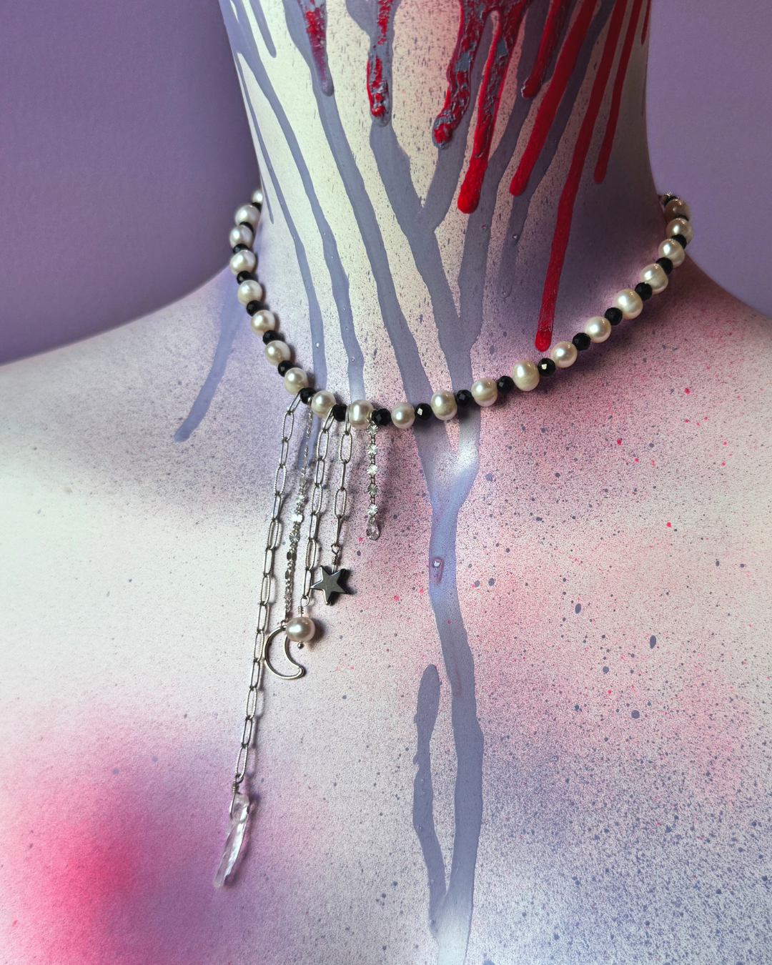 A gothic-style choker necklace displayed on a mannequin torso. The necklace features a strand of black and white pearls, silver chain links, and dangling charms including a silver crescent moon, a silver star, and a silver fish.