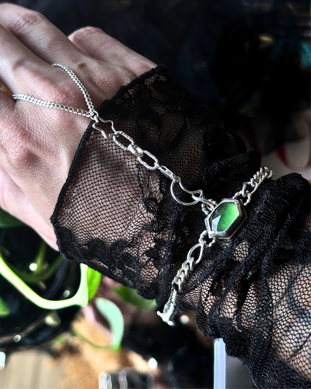 A close-up of a hand wearing a silver chain bracelet with a green faceted gemstone, layered over a black lace wrist cuff.
