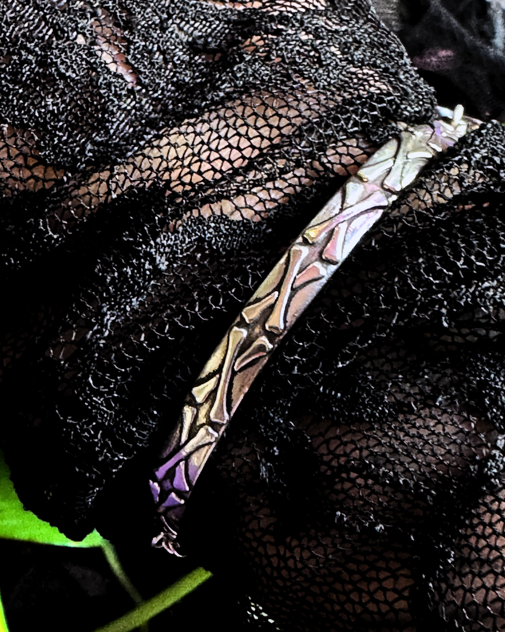 A close-up of a textured metallic bracelet with a geometric, leaf-like pattern, resting on black sheer lace fabric.
