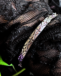 A close-up of a textured metallic bracelet with a geometric, leaf-like pattern, resting on black sheer lace fabric.