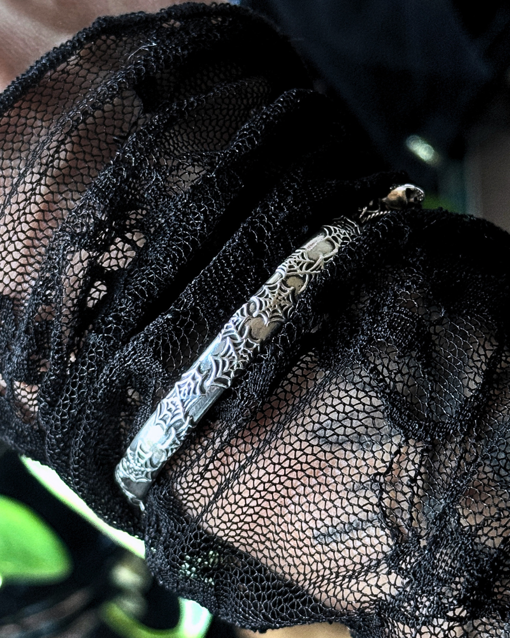 A close-up of a silver-toned bangle bracelet worn on a wrist, partially covered by a sheer black lace cuff. One-of-a-kind sterling silver THE REBIRTH bangle with spider web detailing and skull charm.