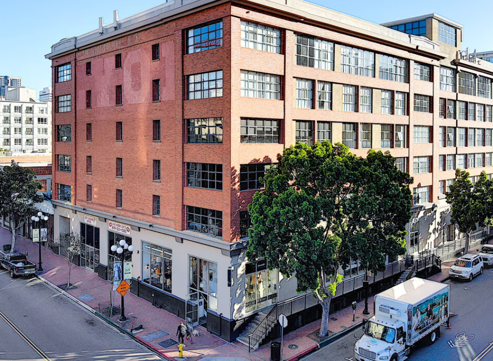 Aerial view of a large brick office building with multiple stories, storefronts on the ground level, trees in front, parked cars, and a white delivery truck on a city street.