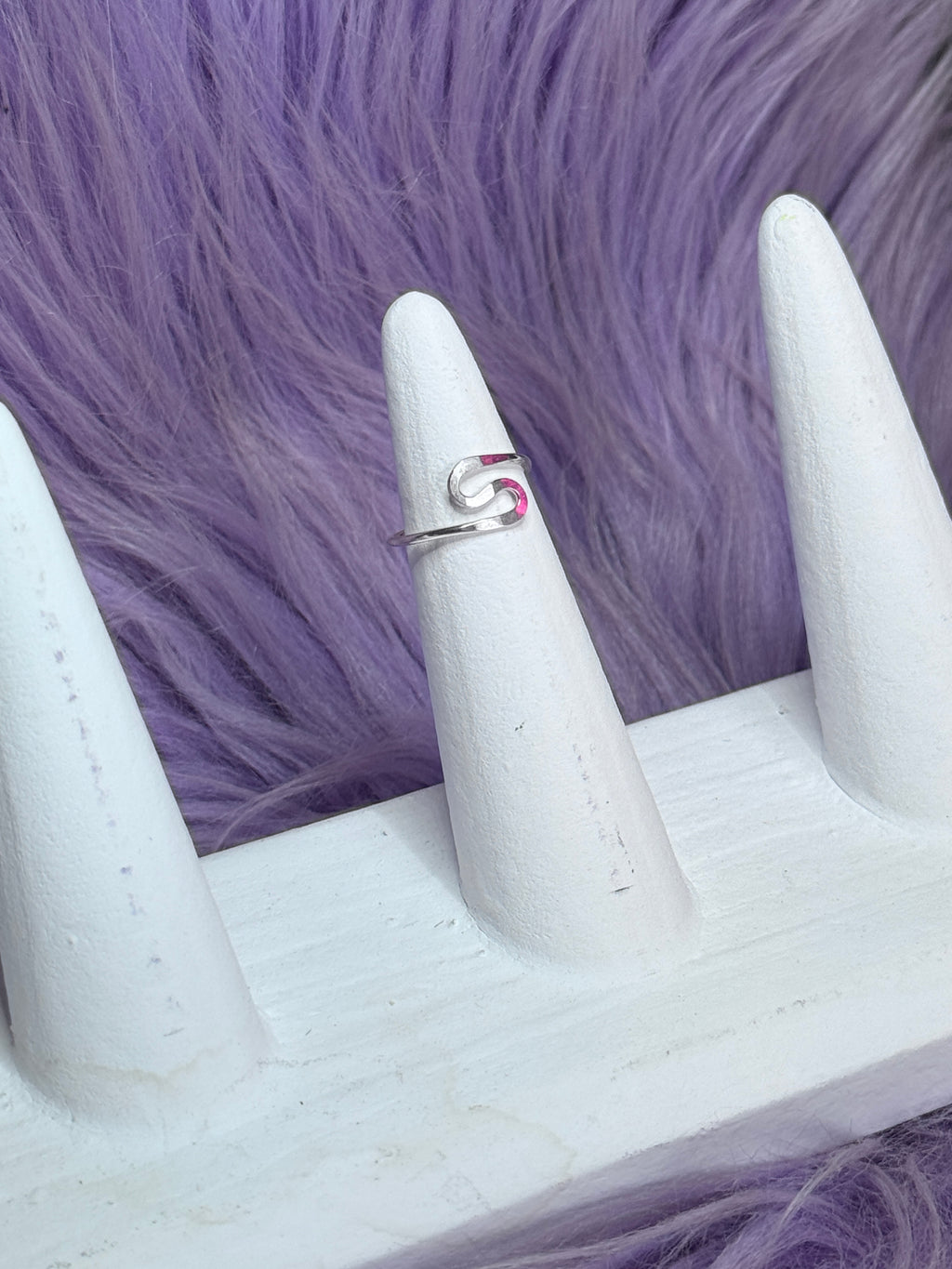 Silver ring with a swirl S shape design on a white stand against a purple fur background