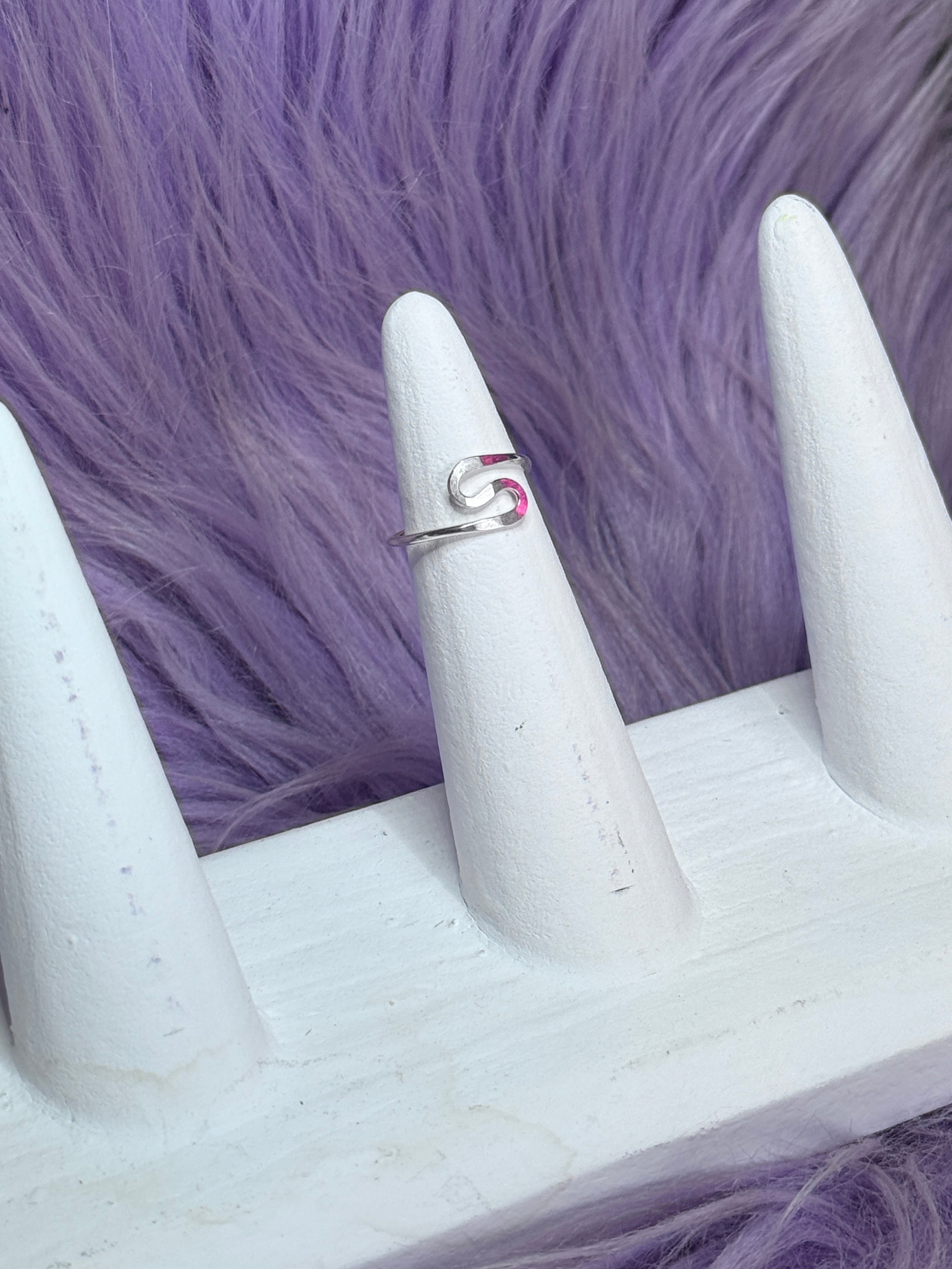 Silver ring with a swirl S shape design on a white stand against a purple fur background