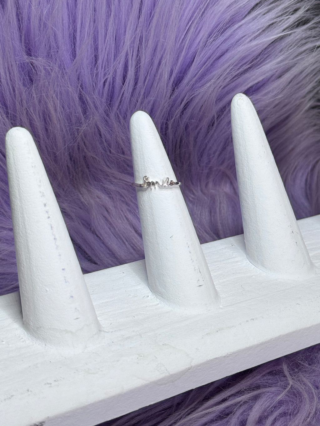 White stiletto nails with a silver ring on a purple fur background and smile written by ring