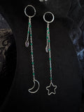 A pair of long silver-toned dangle earrings displayed on a black fabric backdrop. Each earring has a circular hoop at the top, a short chain with green beads, a small faceted crystal charm, and a dangling moon or star-shaped charm.