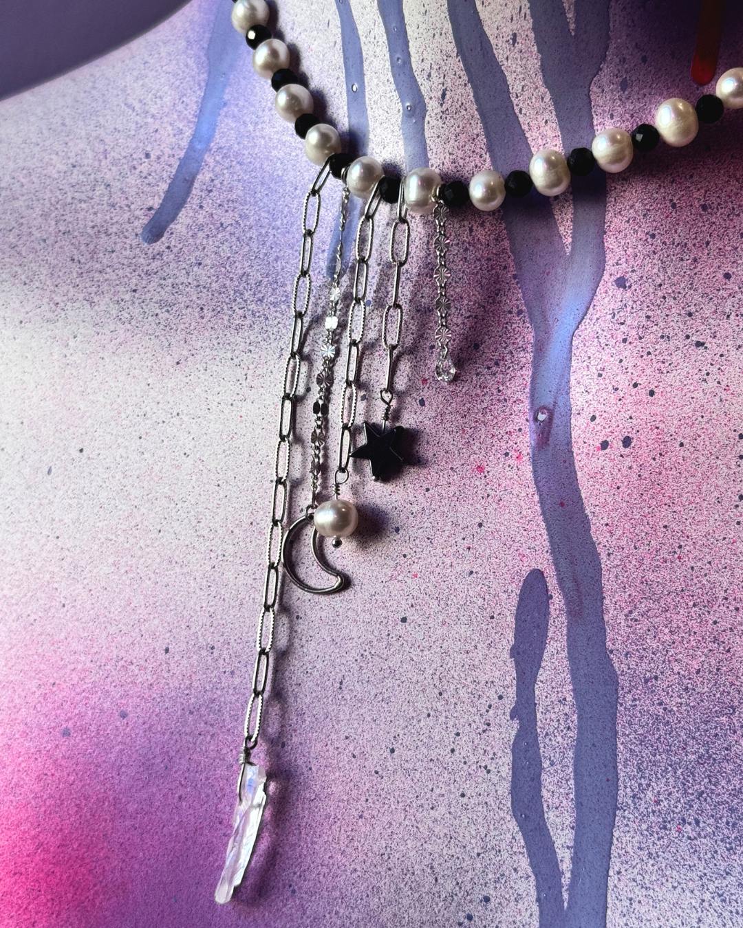A close-up of a person's wrist wearing a silver chain bracelet with a textured, patterned center, styled with a black lace wrist cuff and surrounded by green leaves.