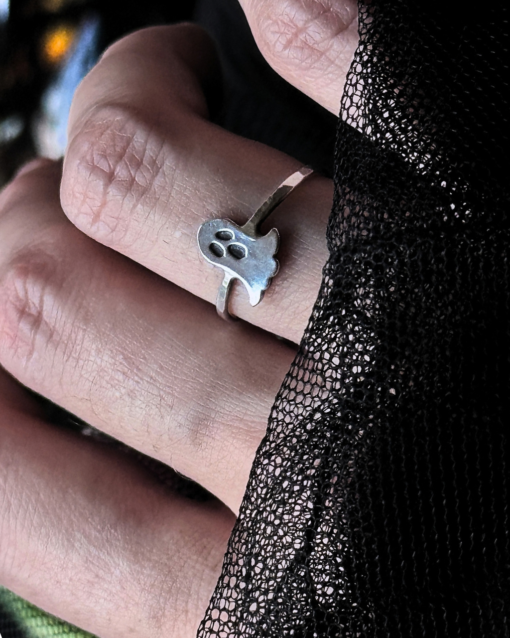 A silver ring shaped like a small ghost with three eye sockets, worn on a person's finger. The hand is framed by a black lace fabric on the right side.