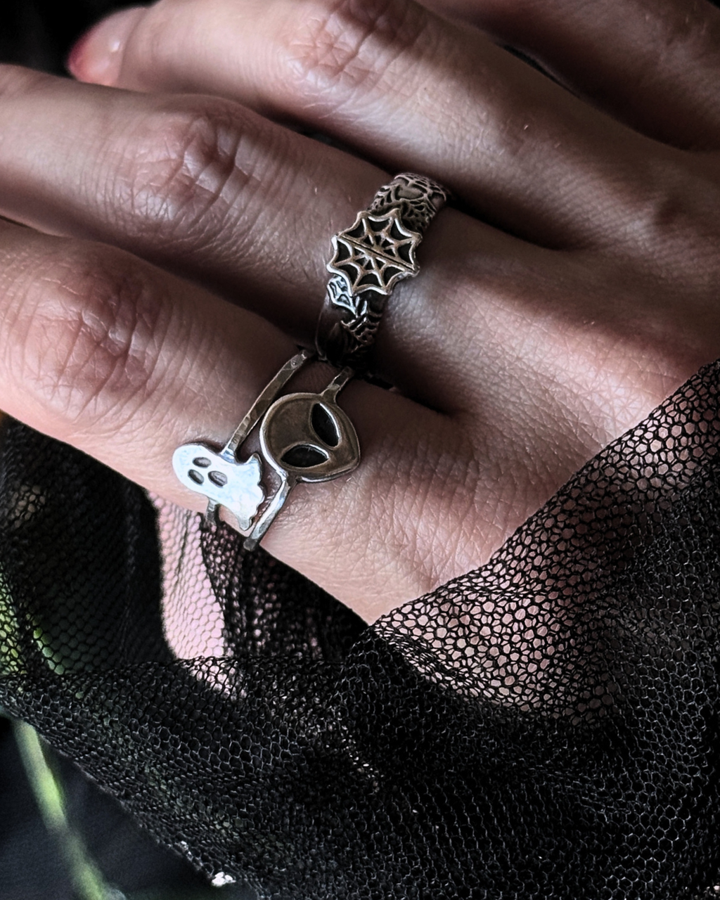 A close-up of a hand wearing three stacked silver rings: one with a spiderweb design, one with a ghost and alien motif, and one with a small metallic charm. The hand is resting on black lace fabric.