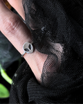 A silver ring with a circular disc face featuring two black oval eyes, worn on a finger and framed by black sheer mesh fabric.