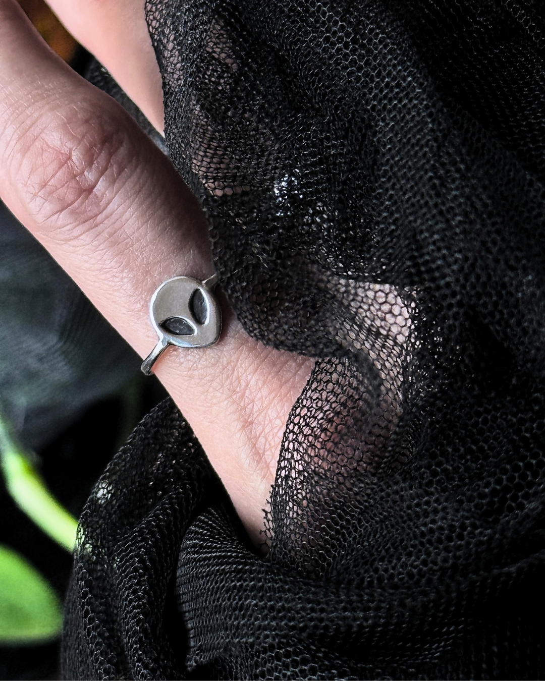 A silver ring with a circular disc face featuring two black oval eyes, worn on a finger and framed by black sheer mesh fabric.