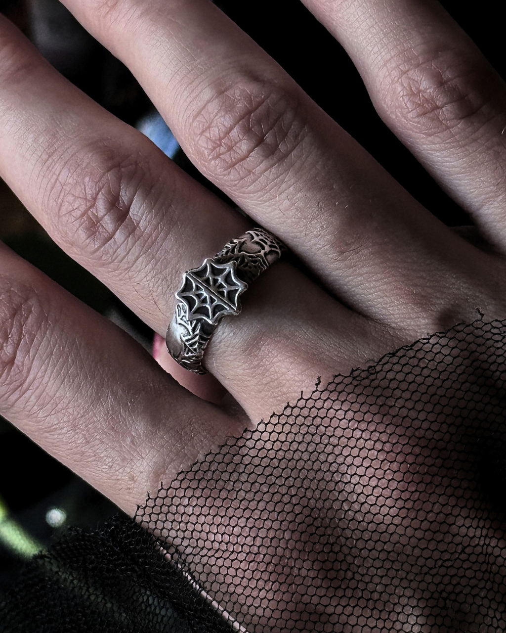 A close-up of a hand wearing a silver ring with an intricate spiderweb design, with black mesh fabric draped over the wrist.