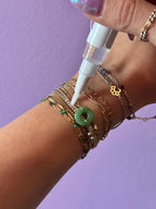 Hand with multiple gold bracelets applying a POLISHING liquid to a green JADE on a purple background