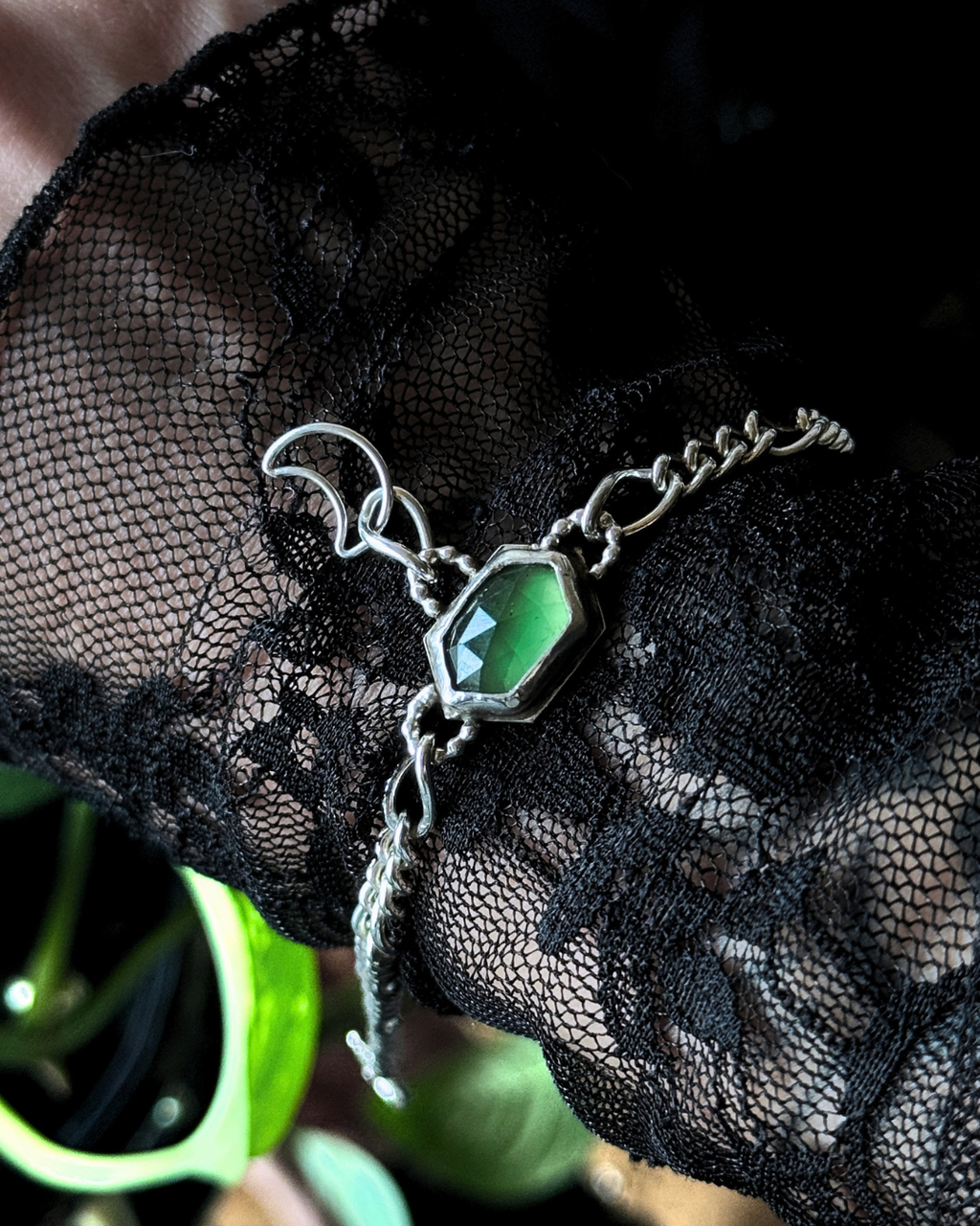 A silver-toned chain bracelet with a faceted green gemstone centerpiece, draped over black lace fabric.