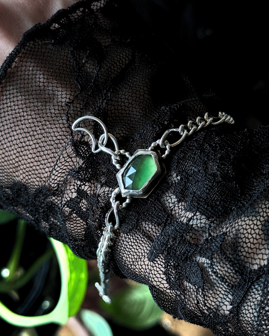 A silver-toned chain bracelet with a faceted green gemstone centerpiece, draped over black lace fabric.