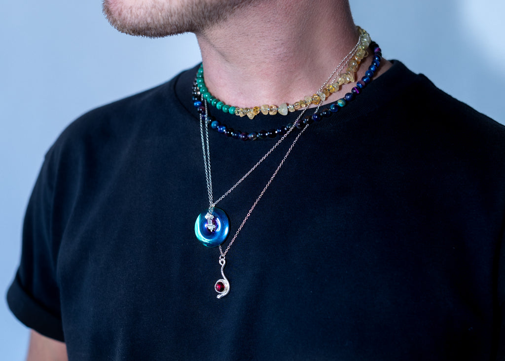Person wearing multiple necklaces with colorful beads and pendants on a black shirt against a light blue background
