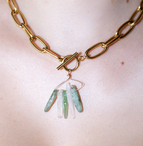 Gold chain necklace with green and clear pendant on a light pink background