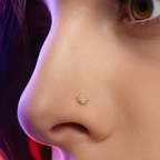 A close-up of a person's nose wearing a small gold nose stud with tiny clear stones, shown against skin with purple and pink lighting.