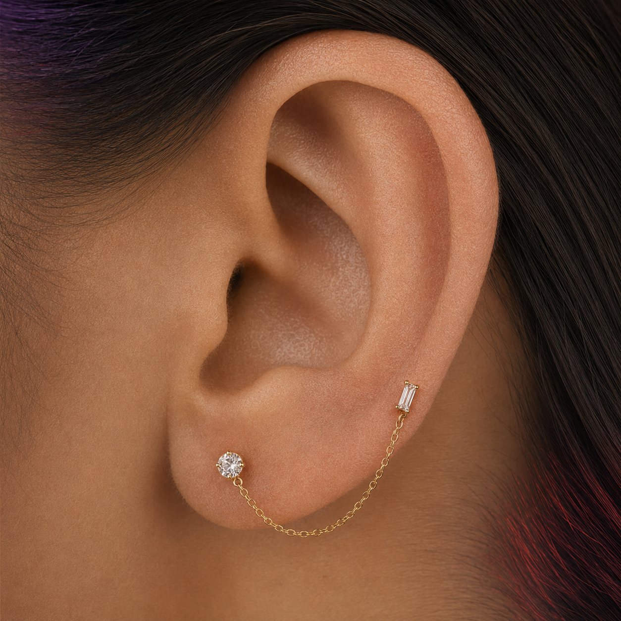 A close-up of a person's ear wearing a delicate gold ear cuff with a chain connecting a small rectangular baguette stud and a round stud, set against dark hair.