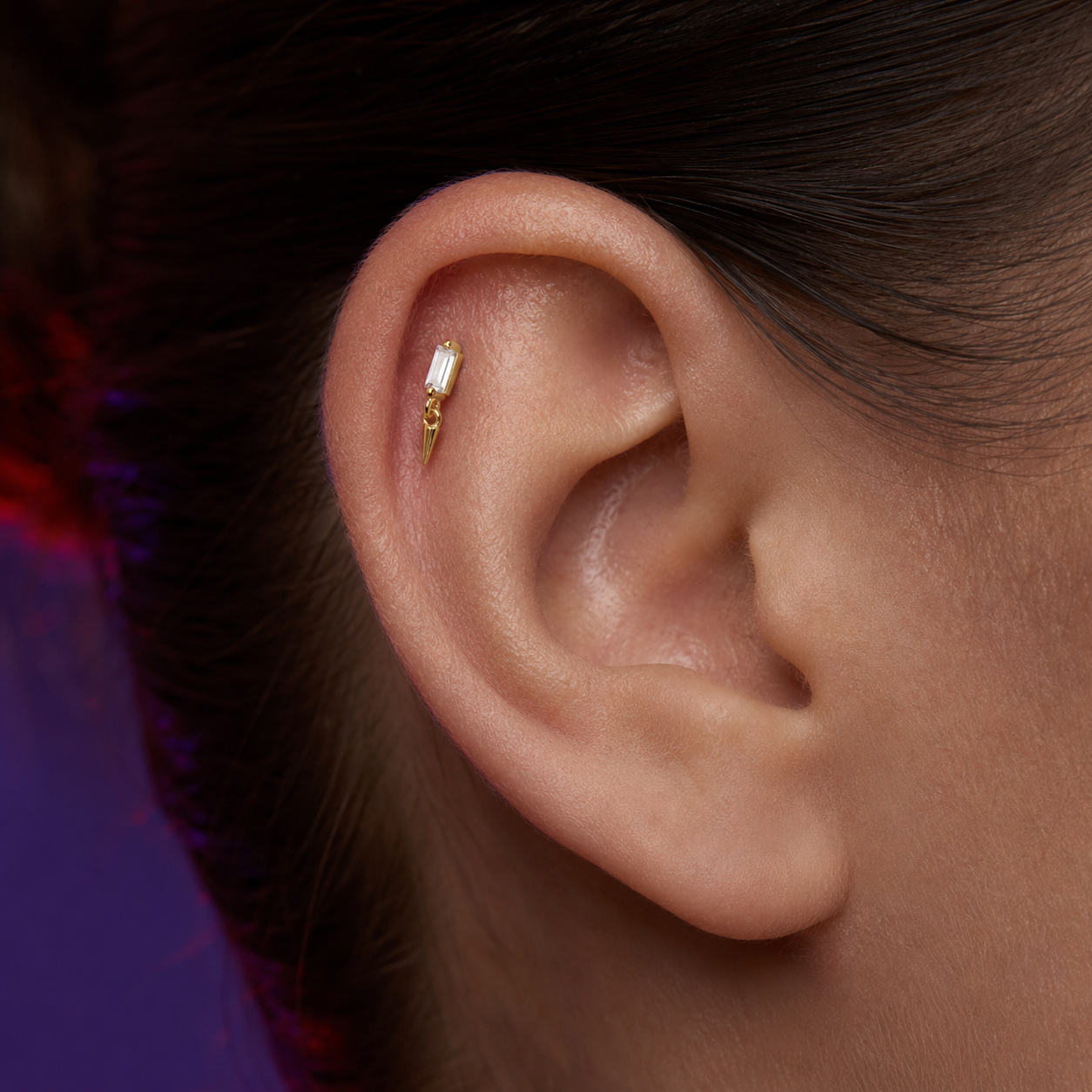 A close-up of a person's ear wearing a delicate gold ear cuff with a small clear rectangular stone, shown against dark hair and a purple-toned background.