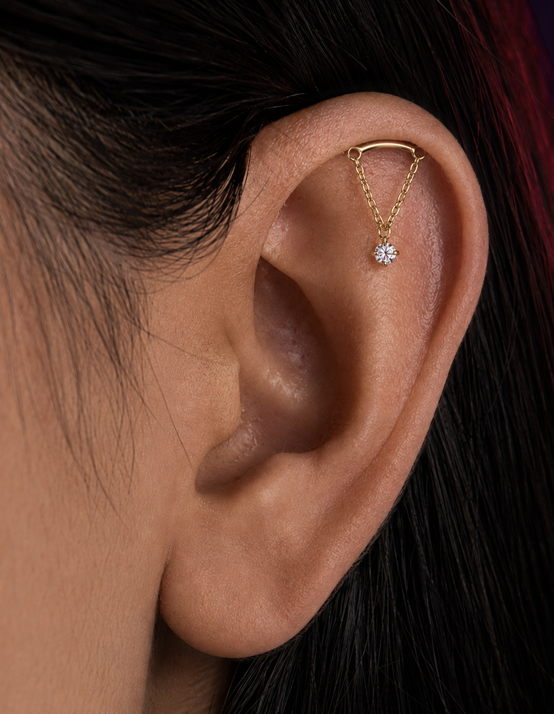 A close-up of a person's ear wearing a delicate gold ear cuff with a small dangling rhinestone charm.