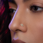 A close-up of a model's nose wearing a small gold nose stud with a halo-like eye design set with clear stones.