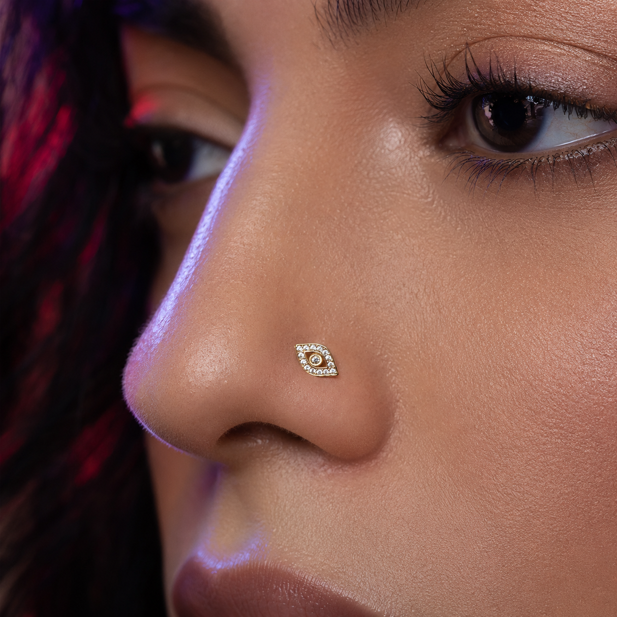 A close-up of a model's nose wearing a small gold nose stud with a halo-like eye design set with clear stones.