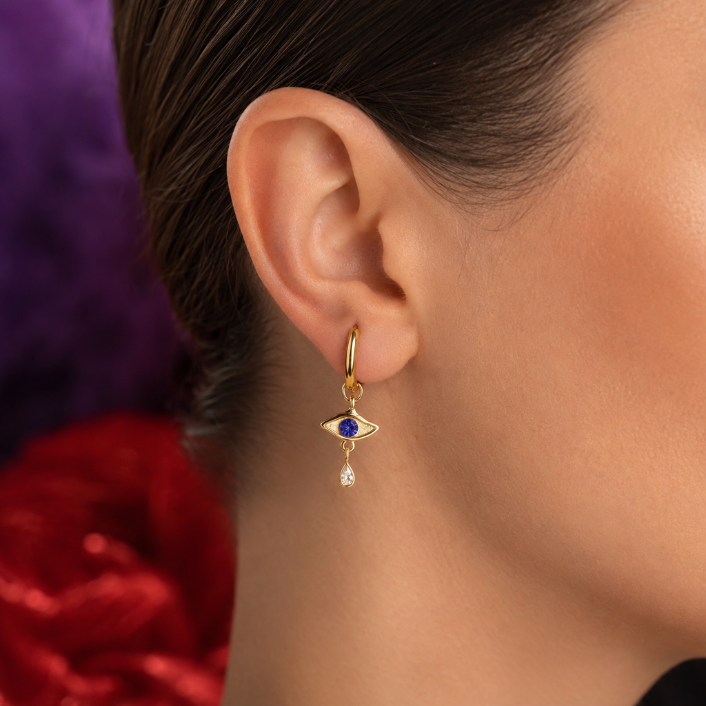 A close-up of a model's ear wearing a gold huggie hoop earring with a dangling evil eye charm featuring a blue stone and a small clear teardrop.