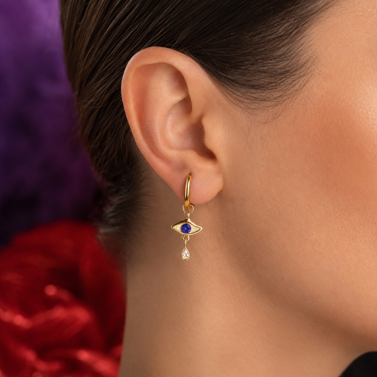 A close-up of a model's ear wearing a gold huggie hoop earring with a dangling evil eye charm featuring a blue stone and a small clear teardrop.