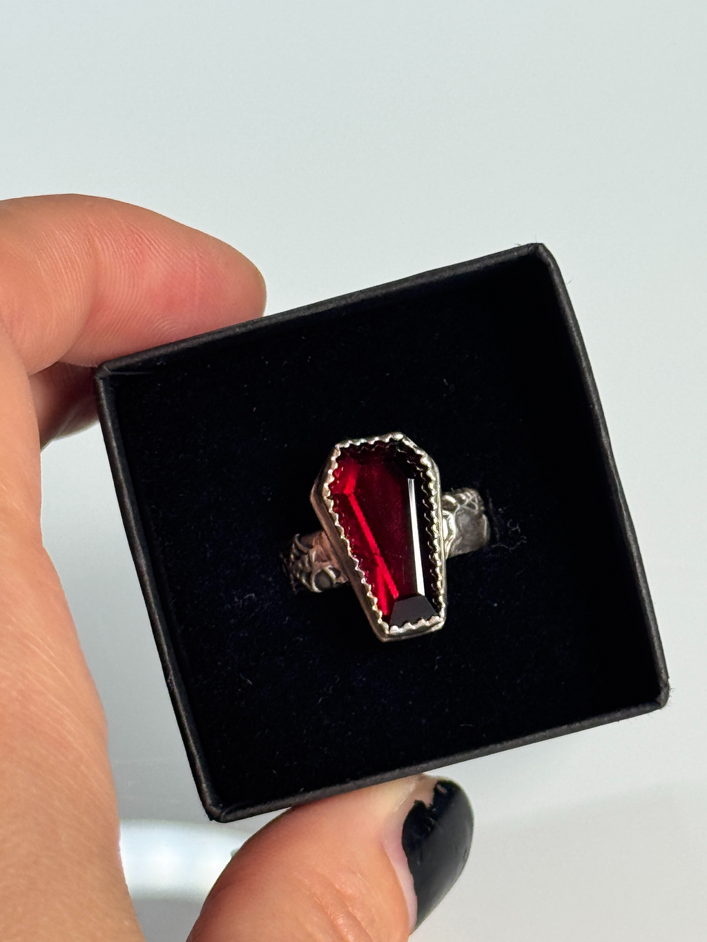 Ring with red stone and silver setting in a black box held by a hand.