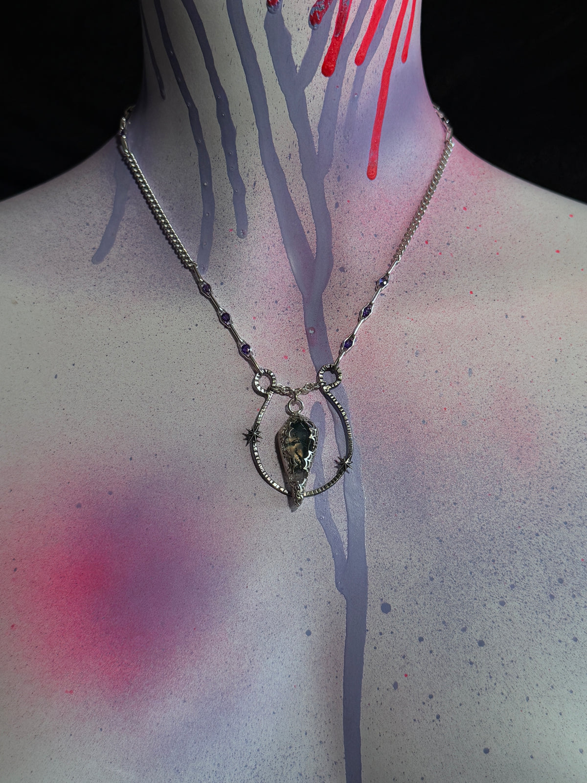 A silver chain necklace with a coffin moss agate pendant and small star charms, displayed on a mannequin torso decorated with spray-paint splatters and drips.