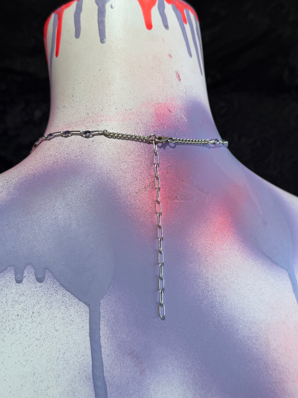 A silver chain necklace with a long drop pendant displayed on the back of a painted mannequin torso. The mannequin has a white base with red, gray, and purple paint drips, set against a dark background.