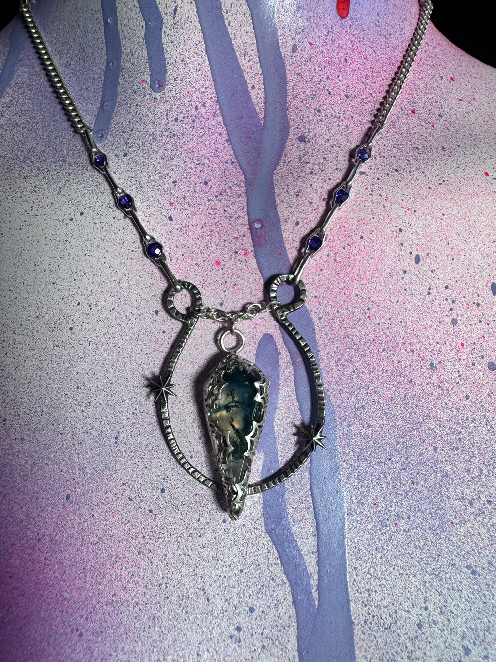 A silver-toned necklace with a large teardrop-shaped moss agate stone pendant, displayed on a mannequin bust against a pink, purple, and blue paint-splattered background.
