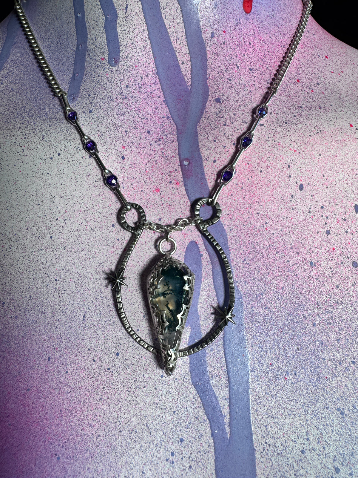 A silver-toned necklace with a large teardrop-shaped moss agate stone pendant, displayed on a mannequin bust against a pink, purple, and blue paint-splattered background.