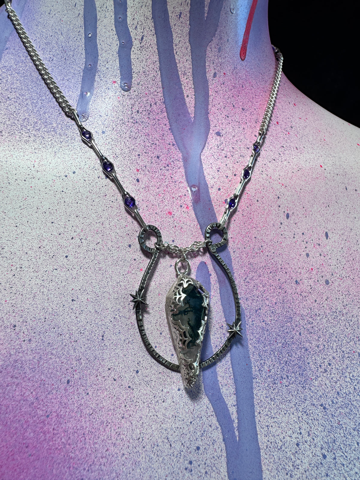 A silver-toned gothic-style pendant necklace displayed on a mannequin neck. The necklace features a central teardrop-shaped dark stone pendant, star-shaped silver charms, and small purple gemstone accents on the chain, set against a purple and blue spray-painted background.