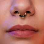 Close-up of a person's nose wearing a gold nose ring with bat wings and a small black heart in the center.