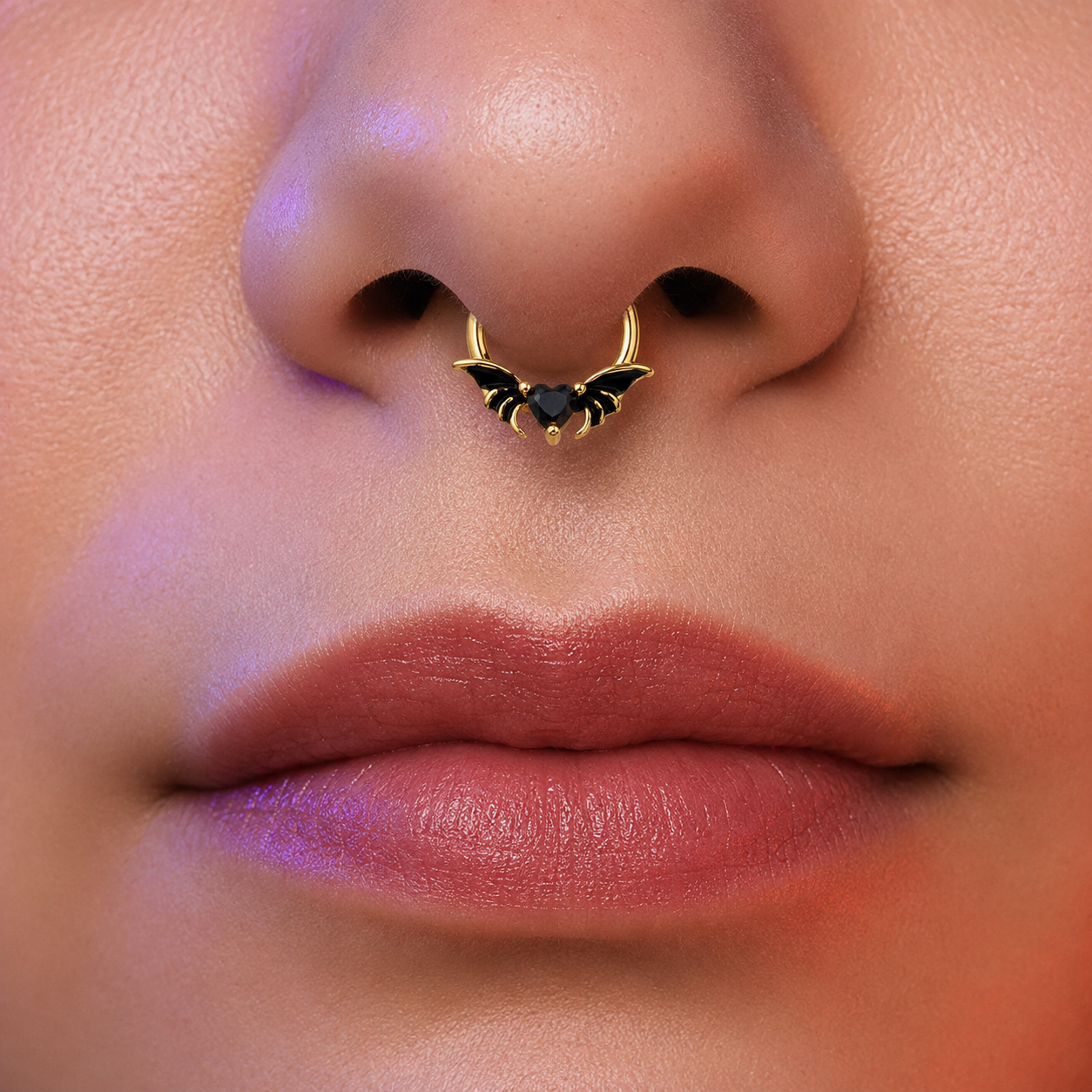 Close-up of a person's nose wearing a gold nose ring with bat wings and a small black heart in the center.