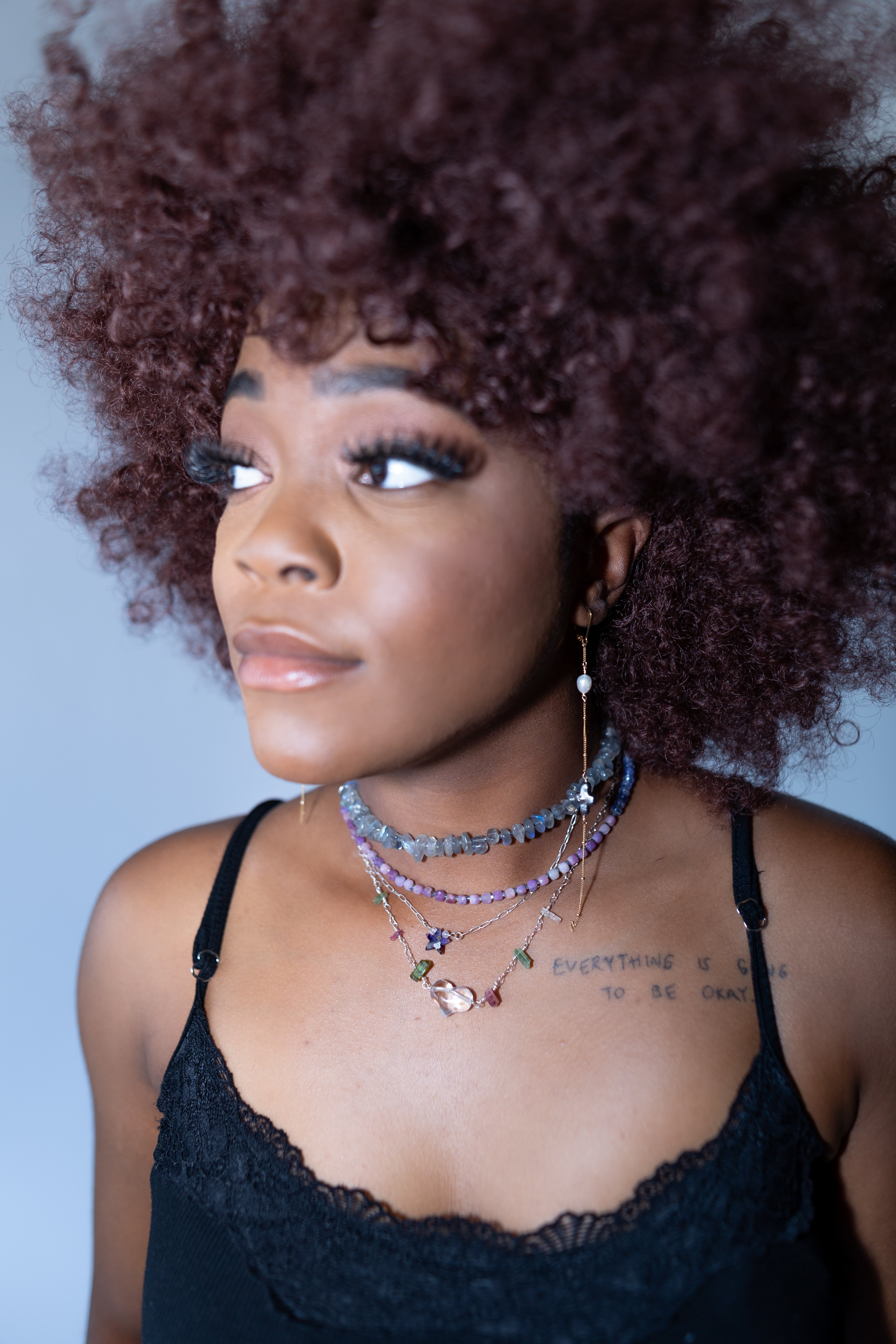 Woman with an afro wearing multiple necklaces against a plain background