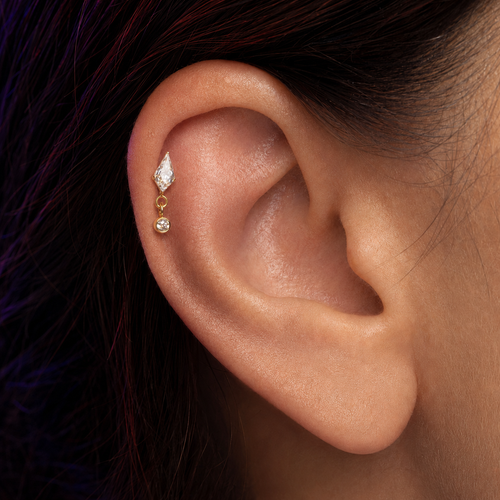 A close-up of a person's ear wearing a gold ear cuff with a marquise-cut clear stone and a small dangling clear stone.