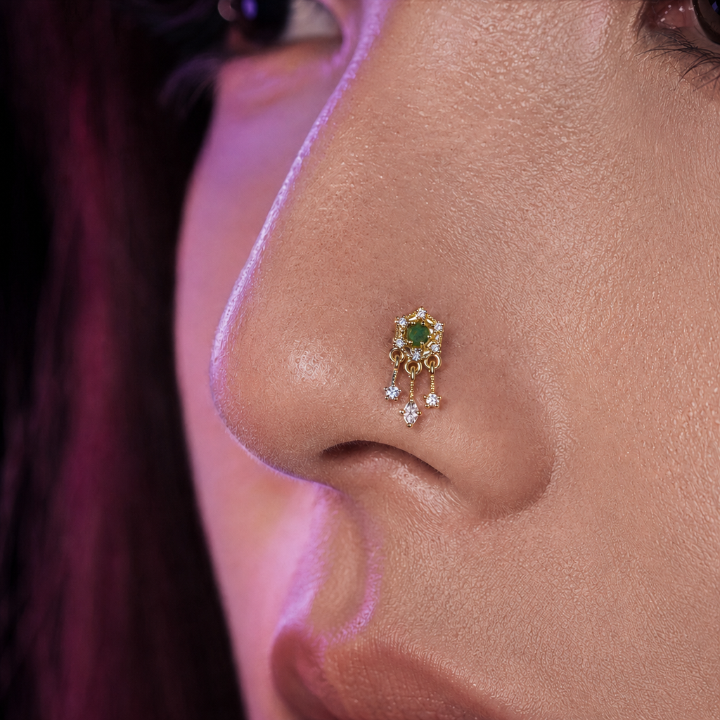 A close-up of a person's nose wearing a small gold nose stud with a green heart-shaped center and dangling clear stones, set against warm skin tones with pinkish lighting.