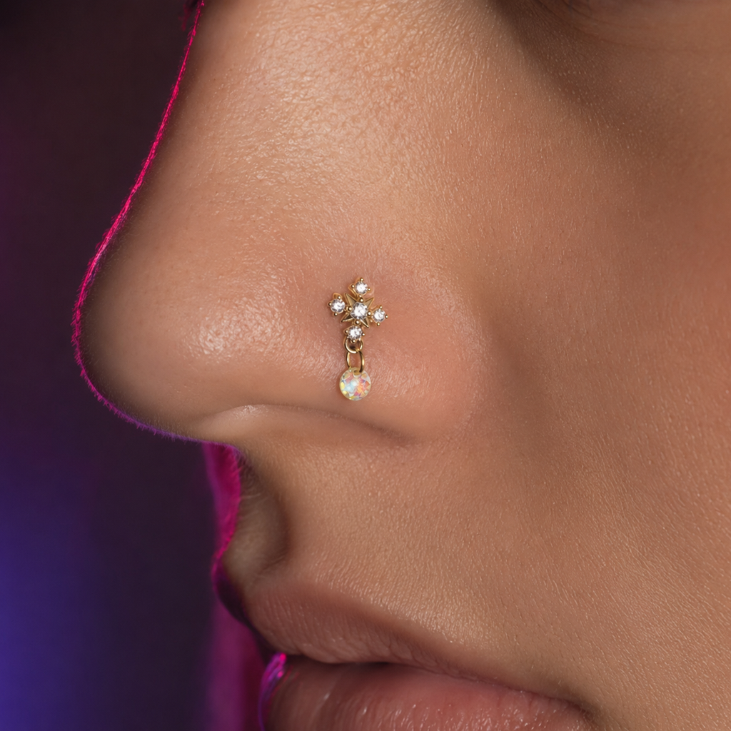 A close-up side profile of a person's nose wearing a small gold nose stud with clear rhinestones and a dangling iridescent pearl-like drop.