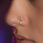 A close-up side profile of a person's nose wearing a small gold nose stud with clear rhinestones and a dangling iridescent pearl-like drop.