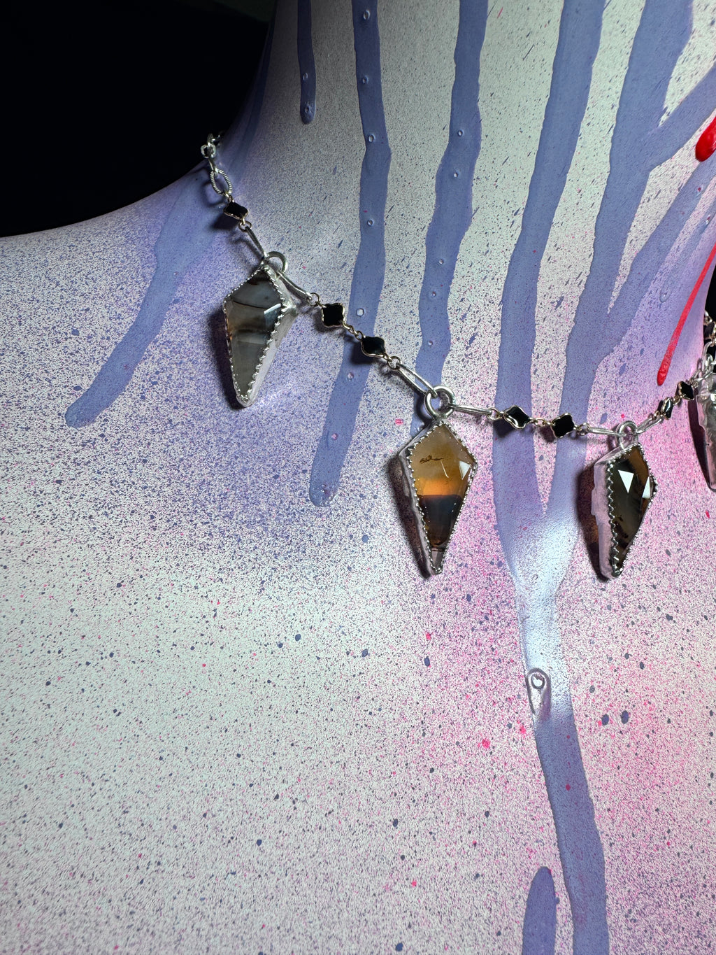 A silver-toned chain necklace with multiple faceted smoky amber-brown geometric pendant charms and small black beads, displayed on a mannequin bust against a paint-splattered background.