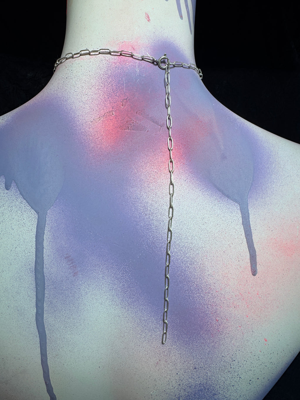 A silver chain necklace with a long dangling link chain and a small clasp, shown on the back of a mannequin or model torso. The torso is painted with a light base and features purple spray-drip effects and pinkish-red splatters against a black background.
