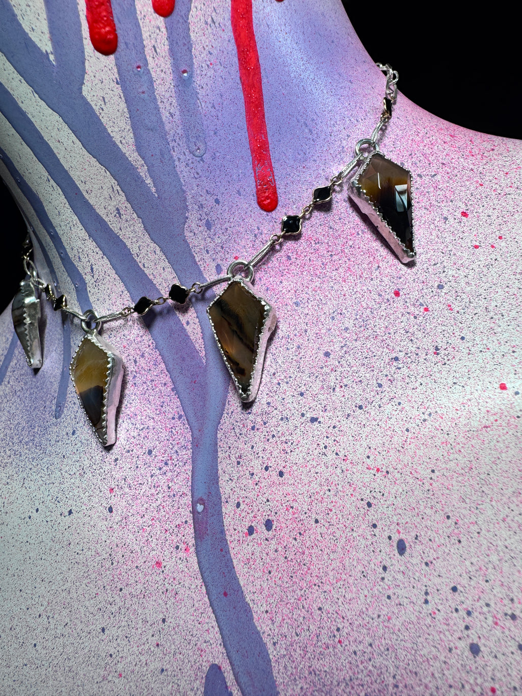 A silver-toned chain necklace with four elongated faceted gemstone pendants, displayed on a painted mannequin torso with pink, purple, and red paint splatter and drips.
